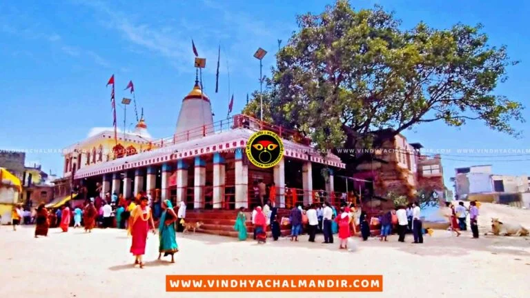 A panoramic view of the Vindhyachal temple complex, a Hindu temple located in Vindhyachal, India. The image shows the temple's entrance with intricate carvings and decorations, as well as several towers and spires. The area around the temple is lush and green, with trees and a river visible in the background.