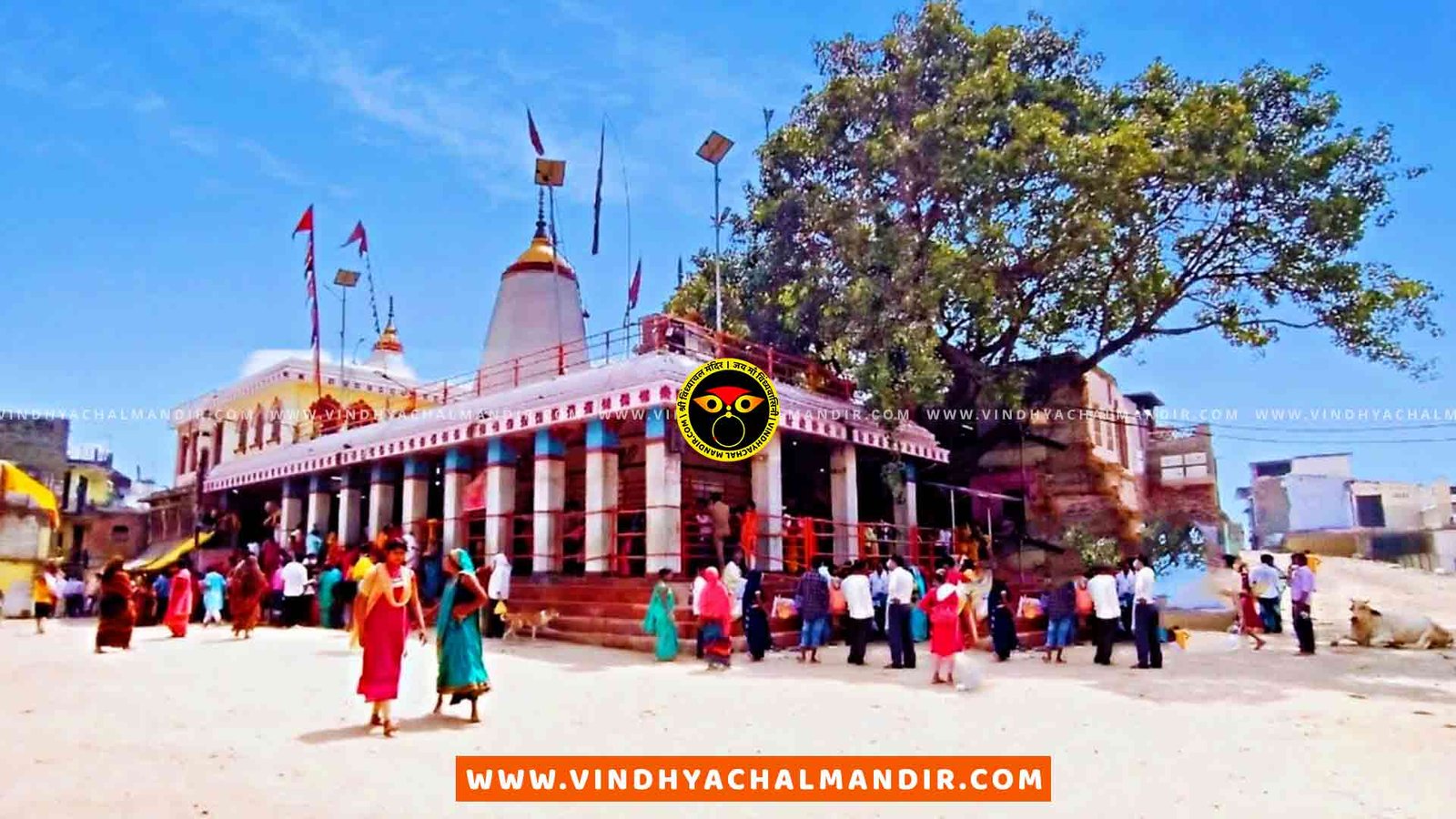 A panoramic view of the Vindhyachal temple complex, a Hindu temple located in Vindhyachal, India. The image shows the temple's entrance with intricate carvings and decorations, as well as several towers and spires. The area around the temple is lush and green, with trees and a river visible in the background.